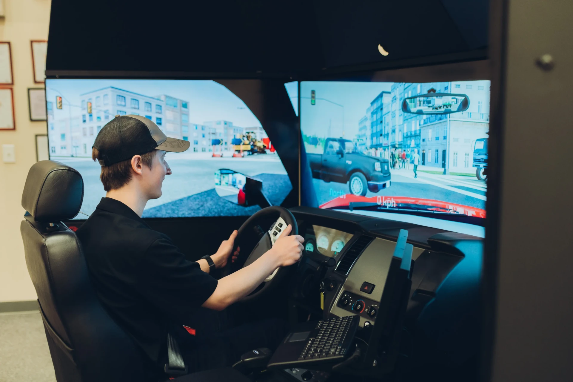 A person wearing a black shirt and cap sits in a driving simulator, holding the steering wheel and looking at large screens displaying a virtual city and road scene.